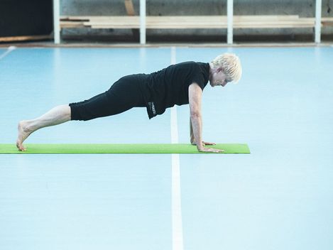 Person mit kurzen hellen Haaren in schwarzer Sportkleidung hält eine Plank-Position auf einer grünen Yogamatte in einer Halle mit blauem Boden
