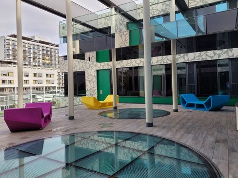 Mehrere farbige Enzos als Sitz- und Liegeelemente in einem modernen, überdachten Innenhof mit Glasflächen und Säulen.