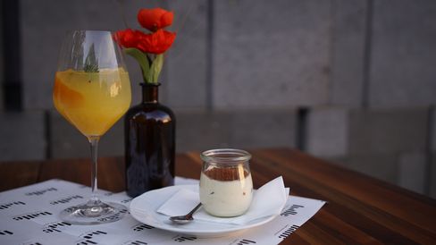 Glass with yellow drink and orange slice, small glass with dessert on white plate, brown vase with red flowers on wooden table
