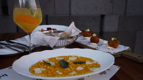 Plate of risotto, wooden board with fried balls on mayonnaise, bread basket with rolls, and glass with orange drink on a table