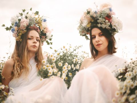 Two women wearing flower crowns sit in a field of daisies wearing white flowing dresses