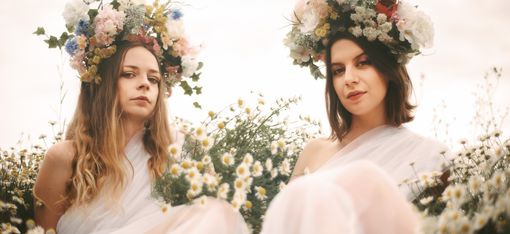 Two women wearing flower crowns sit in a field of daisies wearing white flowing dresses