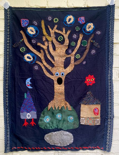 Textile wall hanging showing a large tree with a face, a figure with red feet, a red face, and a small house on a dark background.