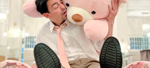 Das Bild zeigt einen Mann, der auf dem Boden sitzt und einen großen rosa Teddybären in den Armen hält. Er scheint die Gesellschaft des Stofftiers zu genießen, das ihn von hinten umarmt.