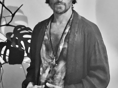 Standing person wearing an open shirt, cardigan, and necklace in front of a plant.