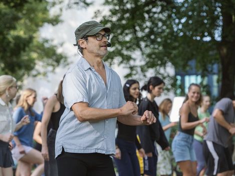 Man wearing glasses and a cap stands outdoors in front of a group of young people who are moving