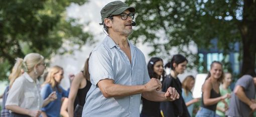Man wearing glasses and a cap stands outdoors in front of a group of young people who are moving