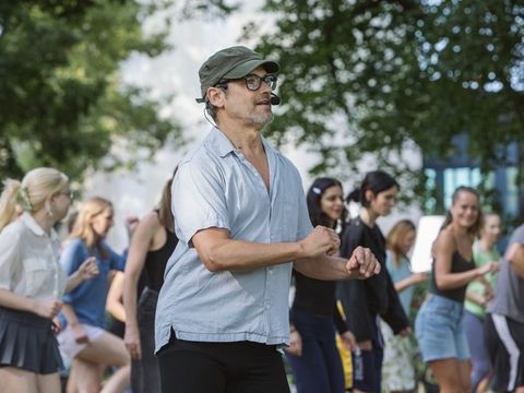 Man wearing glasses and a cap stands outdoors in front of a group of young people who are moving
