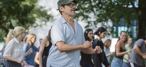 Man wearing glasses and a cap stands outdoors in front of a group of young people who are moving
