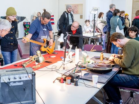 Mehrere Personen in einem Raum beobachten, wie zwei Männer an Gitarren arbeiten, eine steht auf einem Tisch, die andere liegt auf dem Tisch, im Vordergrund ein Verstärker und Werkzeuge