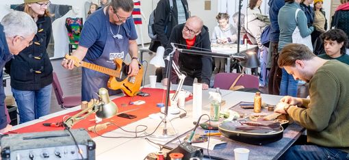 Mehrere Personen in einem Raum beobachten, wie zwei Männer an Gitarren arbeiten, eine steht auf einem Tisch, die andere liegt auf dem Tisch, im Vordergrund ein Verstärker und Werkzeuge