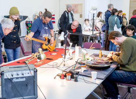 Mehrere Personen in einem Raum beobachten, wie zwei Männer an Gitarren arbeiten, eine steht auf einem Tisch, die andere liegt auf dem Tisch, im Vordergrund ein Verstärker und Werkzeuge
