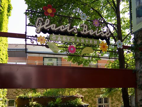Eingangsbereich mit bepflanztem Rankgerüst, darüber ein Schild mit Blumenmotiven und dem Schriftzug 'Glacis Beisl Gastgarten' unter Bäumen.