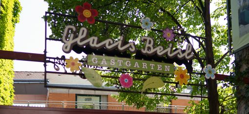 Entrance area with a planted trellis, above it a sign with floral motifs and the words ‘Glacis Beisl Gastgarten’ under trees.