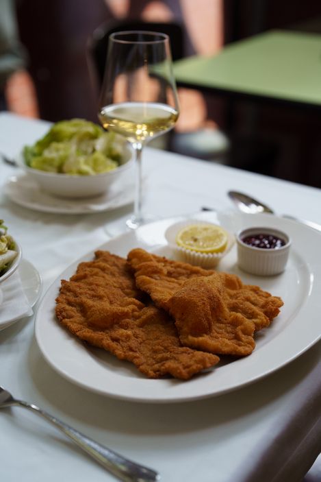 Two breaded schnitzels on a white plate with a slice of lemon, cranberries, and a glass of white wine next to them.