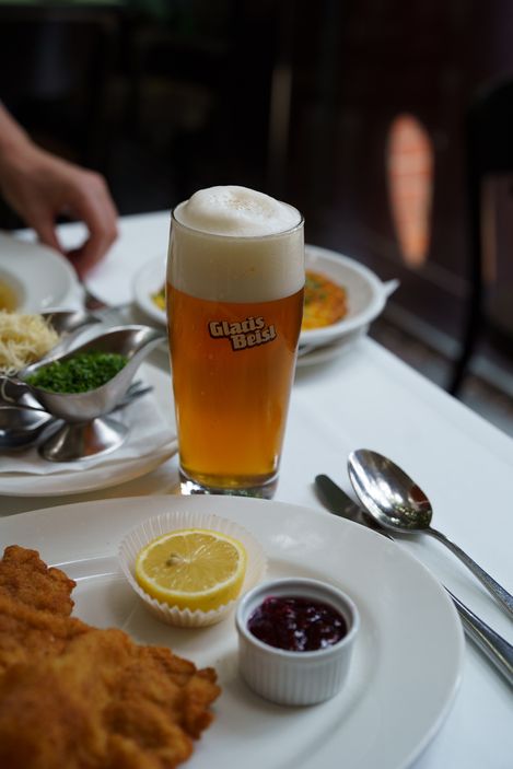 A frothy beer glass next to a plate of schnitzel with a lemon slice and berry sauce. Metal gravy boat with chopped herbs adds a fresh touch.