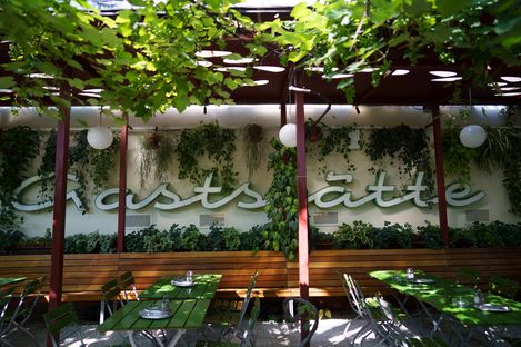 Covered outdoor dining area with wooden benches, green tables, metal chairs, and lush plants on the wall and pergola.