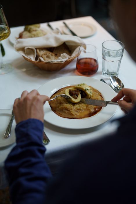 Person cuts a crackling dumpling on a plate with brown sauce, bread basket and drinks on a white-covered table in the background