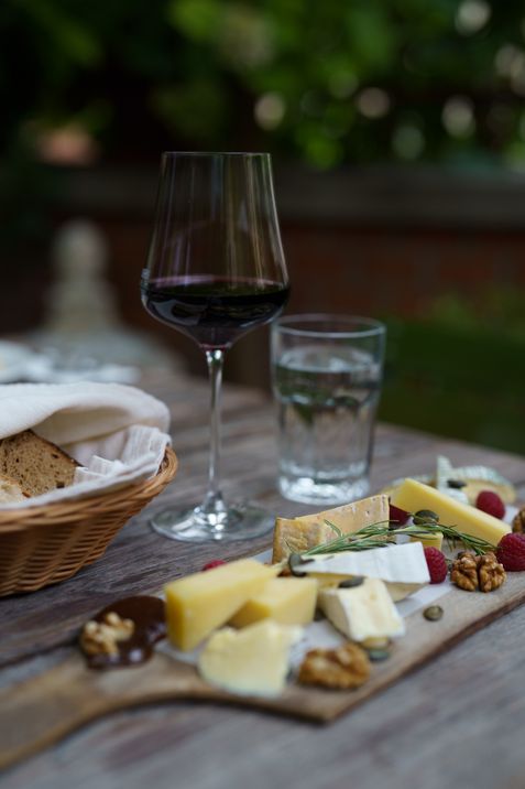 A glass of red wine, a glass of water, a basket of bread, and a wooden board with assorted cheeses, nuts, and raspberries on an outdoor table.