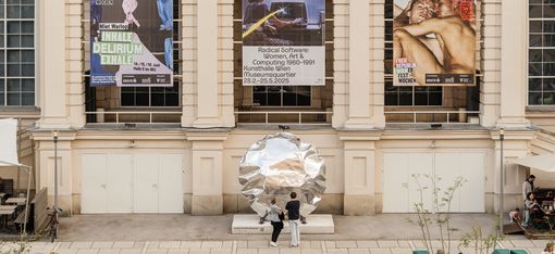 Two people stand in front of a large round shiny metal sculpture against a building facade