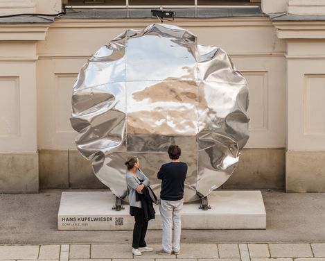 Zwei Personen stehen vor einer großen, runden, glänzenden Metallskulptur vor einer Fassade