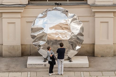 Two people looking at a large silver irregularly shaped sculpture in front of a light-colored wall