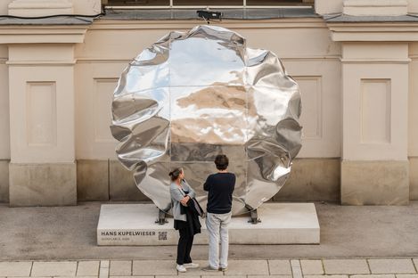 Two people looking at a large silver irregularly shaped sculpture in front of a light-colored wall