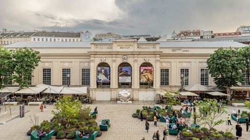 MQ main courtyard with large classical building, in front of it a paved square with trees in planters and seating areas with people.