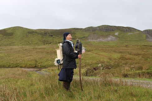 Person with backpack and walking stick standing in open landscape with hills in the background.
