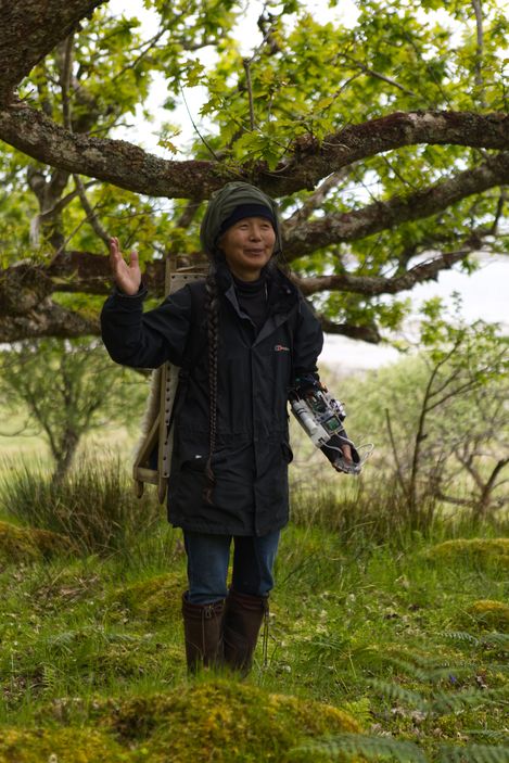Person with long braid wearing a black jacket and rubber boots stands under a tree in a green landscape holding a mechanical device.