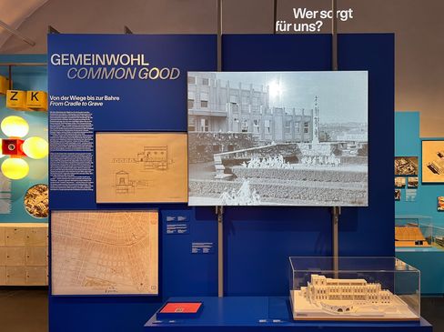 A screen showing a black-and-white film is mounted on a dark blue wall. The words “Gemeinwohl, Common Good” are written on it in large letters. Two drawings and an architectural model are also on display.