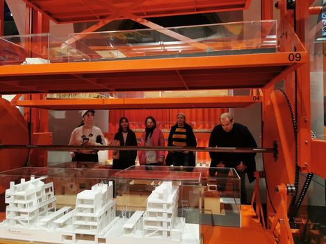 mehrere Personen stehen vor einem großen roten Gerüst auf dem Architekurmodelle stehen. Die Personen schauen sich die Modelle an.