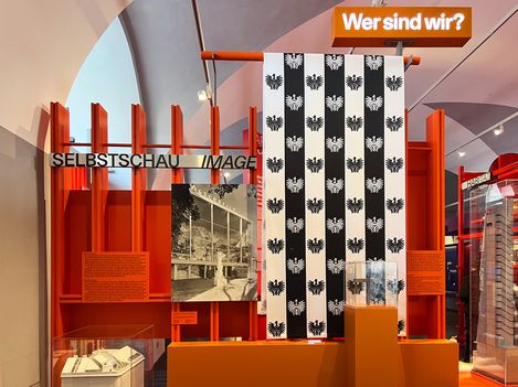 An exhibition wall with black and white stripes on which white and black eagles are printed. Next to it is a photo. Behind it are tall red beams, and above them is the question “Who are we?” in neon letters.