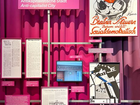 pink-lila Vorhang im Hintergrund, davor einige Fotos und Zeitungsausschnitte. Ein Schild in pink auf dem steht "Antikapitalistische Stadt".