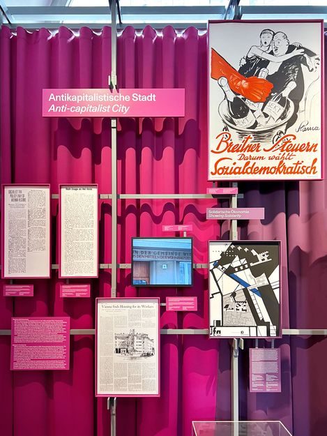 Pink-purple curtain in the background, with several photos and newspaper clippings in front of it. A pink sign that reads “Anti-Capitalist City.”