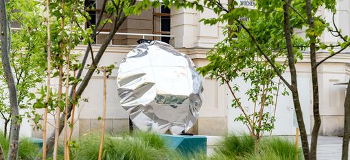 Reflective, irregularly shaped sculpture on a pedestal surrounded by grass and young trees in front of the MuseumsQuartier Vienna