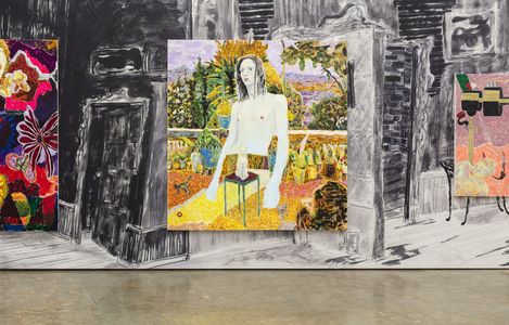 Multiple colorful paintings with abstract and figurative motifs hang in front of a large black-and-white wall drawing.