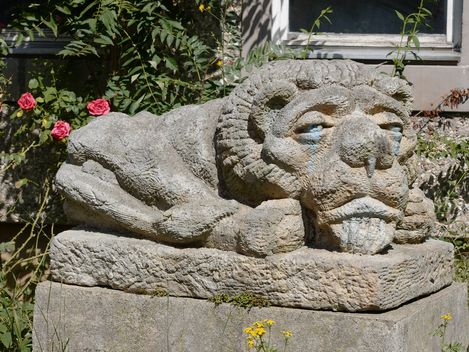 Steinfigur eines liegenden Löwen mit menschlich wirkendem Gesicht in einem Garten, umgeben von Pflanzen und Blumen.