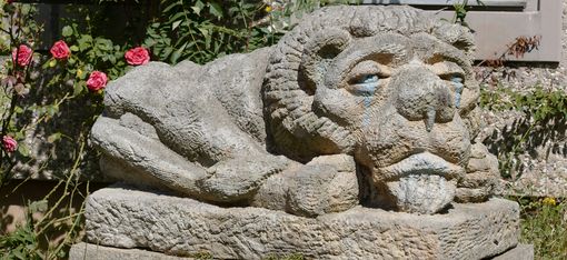 Steinfigur eines liegenden Löwen mit menschlich wirkendem Gesicht in einem Garten, umgeben von Pflanzen und Blumen.