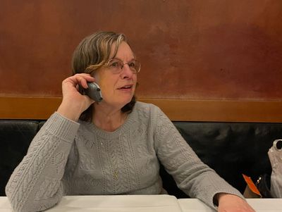 A person with short gray hair and gray knitted sweater sitting at a table holding a phone to their ear.