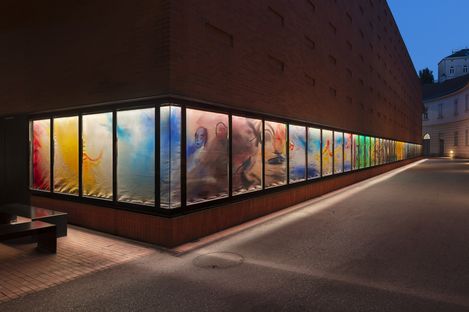 Fassade eines modernen Backsteingebäudes bei Nacht mit einer langen, beleuchteten Fensterinstallation, die abstrakte, farbenfrohe Kunstwerke und figurative Elemente zeigt.