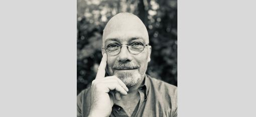 Portrait of a smiling bald man wearing glasses with his index finger resting on his right cheek.