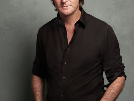 Man wearing a straw hat and black shirt standing against a gray background with hands in pockets.