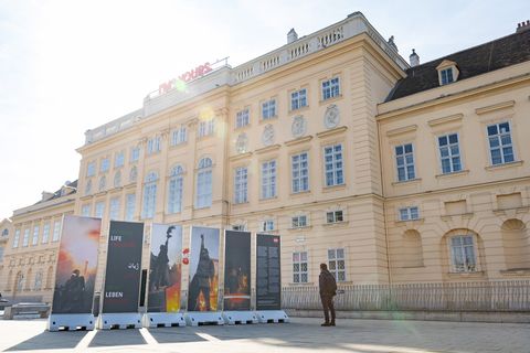 Several large upright posters with images and text in front of a historic light-colored building with many windows and a man observing them