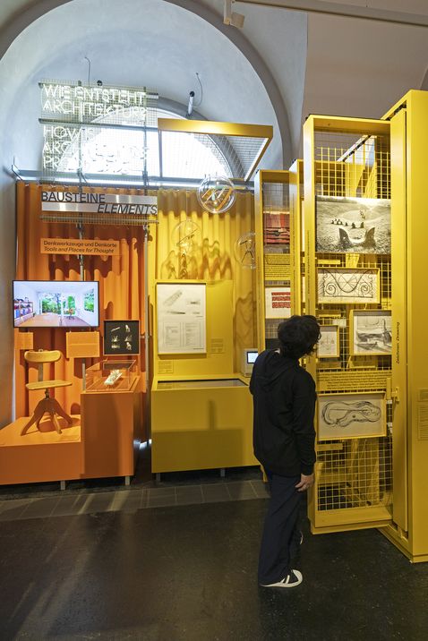 A person dressed in dark clothing looks at pictures and drawings mounted on a yellow grid. Further up, there is a sign that reads “How is architecture created?”