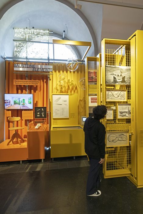 A person dressed in dark clothing looks at pictures and drawings mounted on a yellow grid. Further up, there is a sign that reads “How is architecture created?”