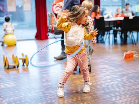 Das Bild zeigt ein junges Kind, das einen Hula-Hoop-Reifen um die Hüften kreisen lässt. Es steht mitten in einem hellen, offenen Innenraum mit Holzfußboden. Im Hintergrund sind weitere Kinder und Erwachsene zu sehen.