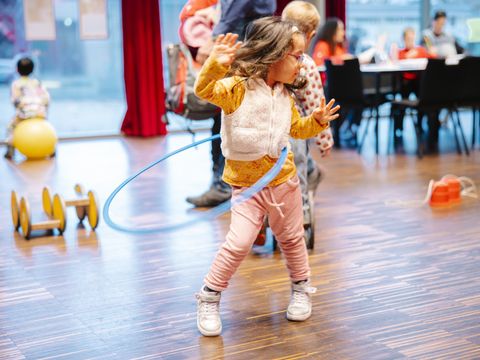 Das Bild zeigt ein junges Kind, das einen Hula-Hoop-Reifen um die Hüften kreisen lässt. Es steht mitten in einem hellen, offenen Innenraum mit Holzfußboden. Im Hintergrund sind weitere Kinder und Erwachsene zu sehen.