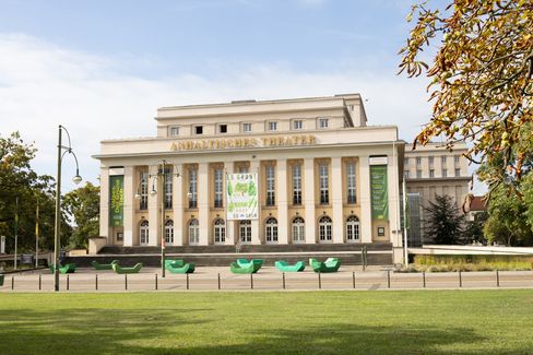 Neoklassizistisches Gebäude mit der Aufschrift „Anhaltisches Theater“, gekennzeichnet durch große Fenster und Säulen. Grüne Banner, Bänke und umgebende Bäume vermitteln eine lebendige Atmosphäre