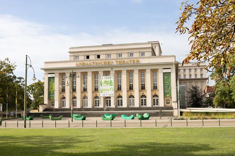 Neoklassizistisches Gebäude mit der Aufschrift „Anhaltisches Theater“, gekennzeichnet durch große Fenster und Säulen. Grüne Banner, Bänke und umgebende Bäume vermitteln eine lebendige Atmosphäre